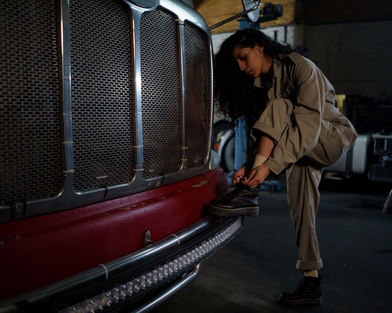 services-01 A woman mechanic in coveralls ties her shoes near a truck bumper in a garage setting.
