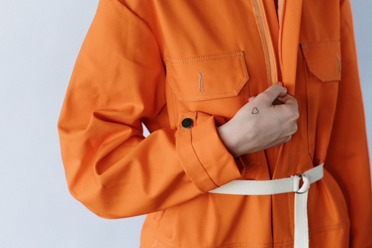 hero-img-02 Close-up photo of a person in a stylish orange jumpsuit, highlighting a small heart tattoo on their hand.