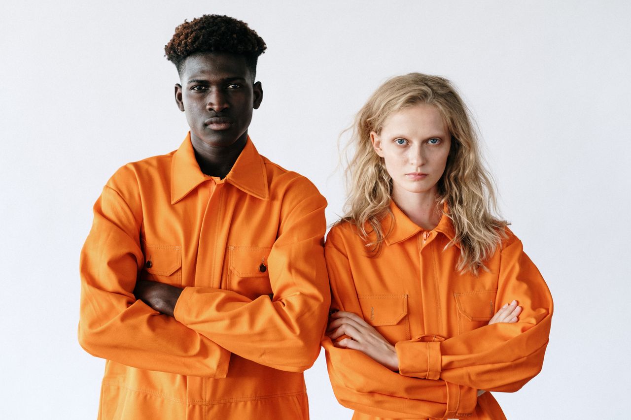 gallery-6 Portrait of a diverse pair in vibrant orange work uniforms with determined expressions.